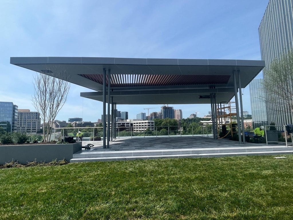 A modern open-air pavilion with a metal roof and columns is under construction in an urban park. Workers wearing safety gear can be seen at the site, handling sheet metal. The pavilion overlooks a cityscape with buildings and greenery in the background. The sky is clear.