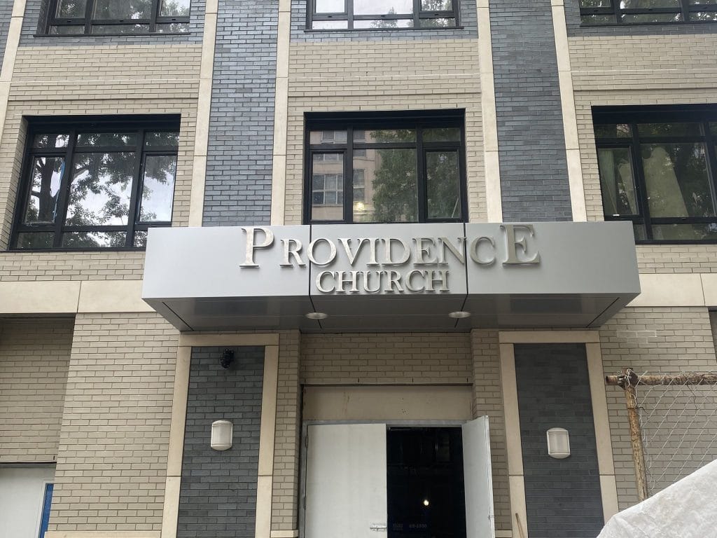 The building facade features a sign reading "Providence Church." The sign is mounted above an entryway with an open door. The exterior wall boasts fiber cement cladding and a mix of light and dark bricks, with windows reflecting the surrounding trees and buildings.