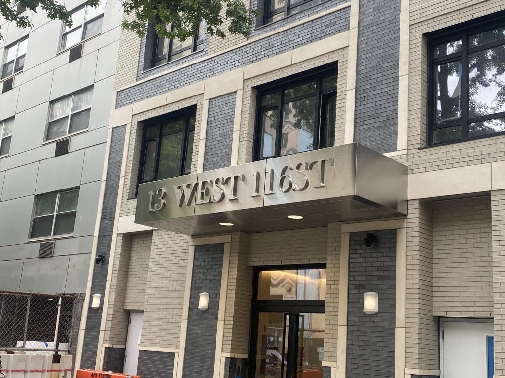 The image shows the entrance of a building with an address displayed as "13 West 116th." The building features a modern exterior facade cladding with grey bricks and large windows. Trees and construction barriers are visible nearby.