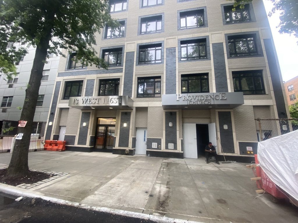 A street view of a multi-story building with the address "18 West 165th St" and a sign for "Providence" on the ground floor. The entrance features large glass doors framed with sheet metal, and there is a person sitting outside near the entrance. Trees line the sidewalk.