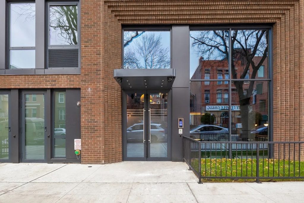 A modern building entrance with large glass doors and windows framed by brick walls. A metal awning covers the doorway. Reflections of nearby buildings and trees are visible in the glass. Fiber cement exterior facade cladding adds a contemporary touch, while a small patch of lawn with a black railing is to the right.
