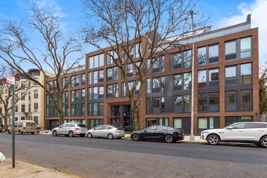 A modern four-story apartment building with large windows and a mix of dark and light brick exterior features sleek standing seam accents. Six cars are parked along the street in front. Leafless trees are on the sidewalk, and the sky is clear with a few clouds.