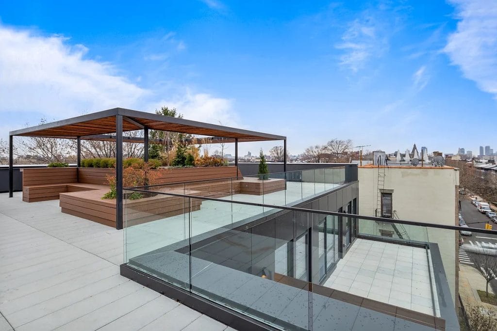 A spacious rooftop terrace features modern glass railing, wooden seating areas, lush greenery, and a pergola providing shade. The city skyline and buildings with exterior facade cladding are visible in the background under a bright blue sky with scattered clouds.