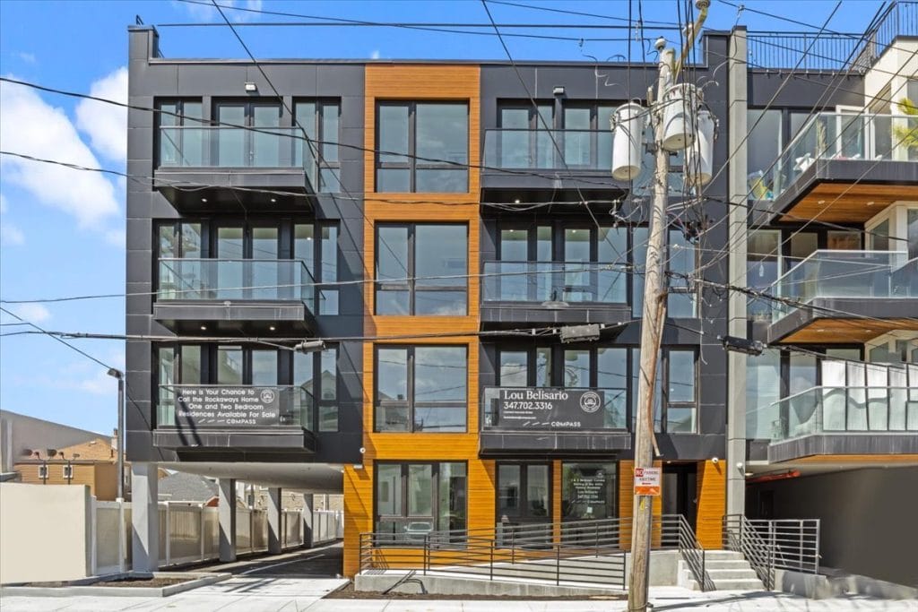 A contemporary apartment building with an Exterior Facade Cladding featuring large windows, glass balconies, and wood accents. Several power lines and a utility pole are visible in front of the building. A sign on the building advertises available units with contact information.
