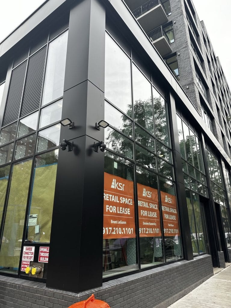 A modern building with large glass windows and black frames features "Retail Space for Lease" signs from KSR. The signs display contact information. The sidewalk adjacent to the building is empty, and there's minimal reflection on the glass, enhancing its sleek appearance.