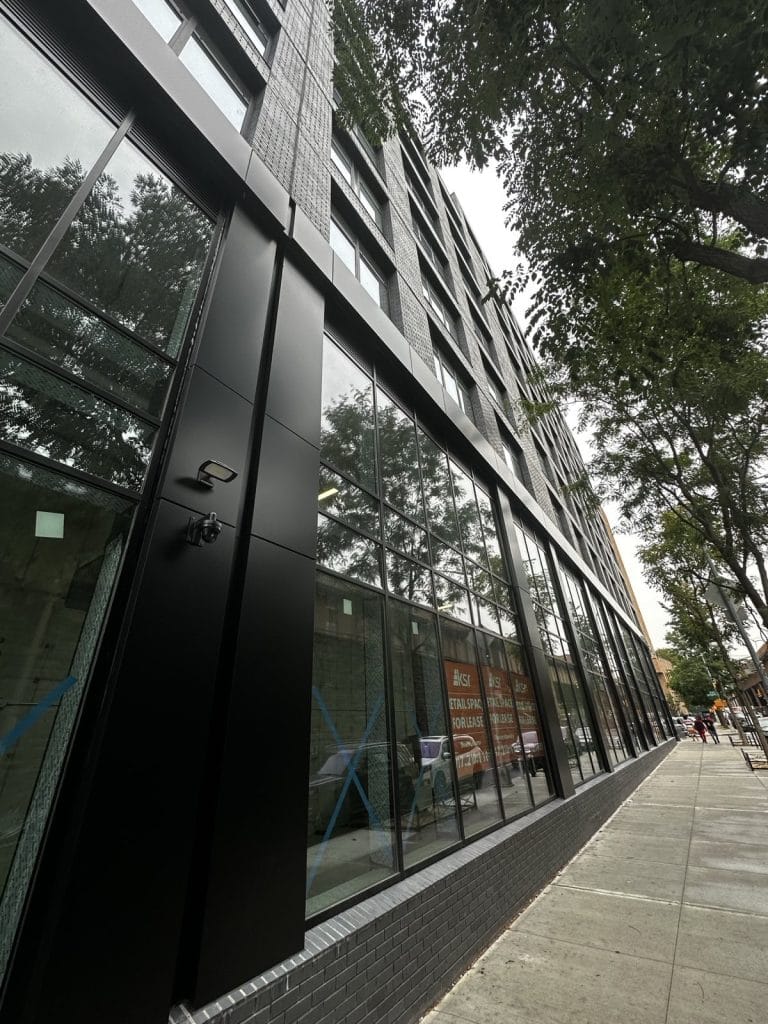 A tall, modern building with large reflective glass windows stretches up alongside a city sidewalk lined with trees. The structure features sleek black panels and a sign in one window advertising leasing opportunities for office space.