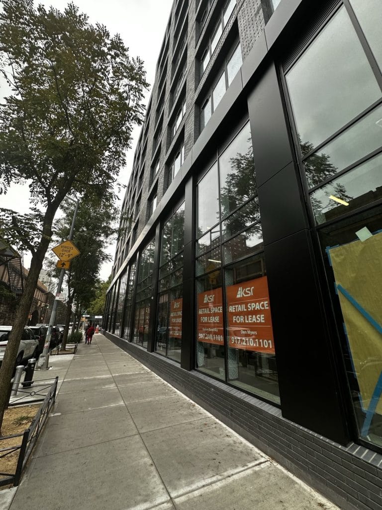 A modern glass-fronted building with several "Retail Space for Lease" signs visible in the windows. The sidewalk adjacent to the building is lined with trees, a bench, and parking meters. A person can be seen walking away in the distance on the sidewalk, adding a touch of life to the scene.