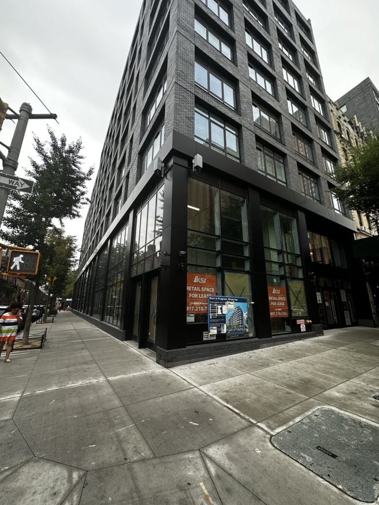 A street view of a multi-story building with large glass windows on the lower level and a dark exterior facade. Signs in the windows advertise retail space for lease. The clean sidewalk, lined with trees, features a pedestrian crosswalk.