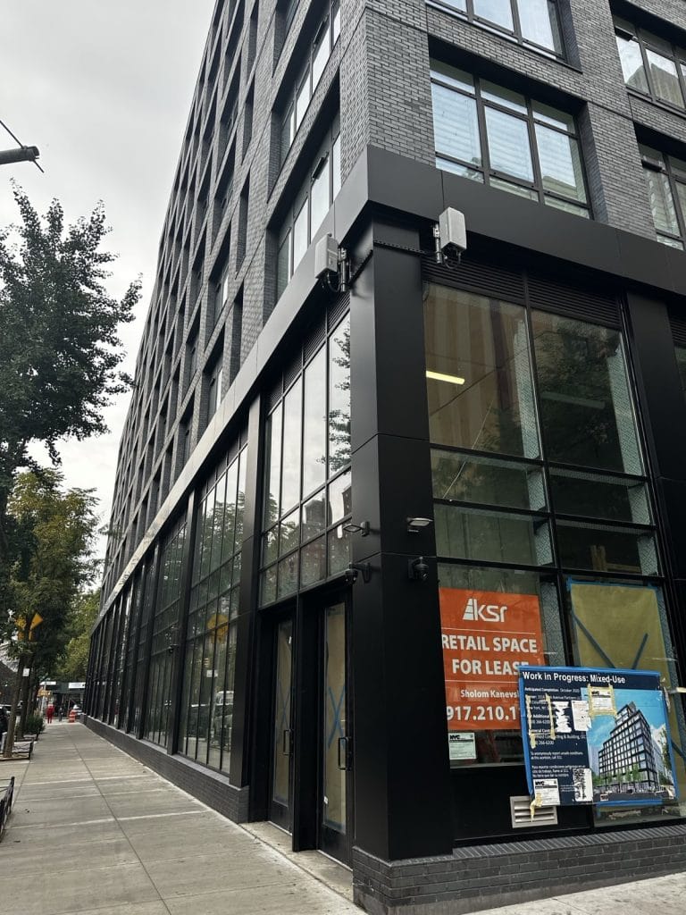 A modern, multi-story building with large windows stands on a city street. The ground floor features a retail space for lease with an orange sign displaying contact information. A tree-lined sidewalk and a streetlight with a traffic signal complete the urban scene.