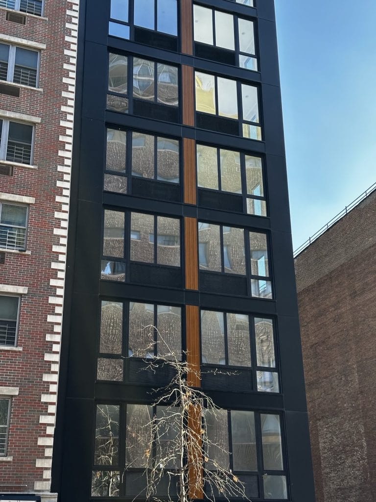 A sleek, tall, modern building with a narrow black facade and large windows stands between two brick structures, one noticeably red. A leafless tree stretches in front of the edifice, juxtaposed against the sky visible in the background.