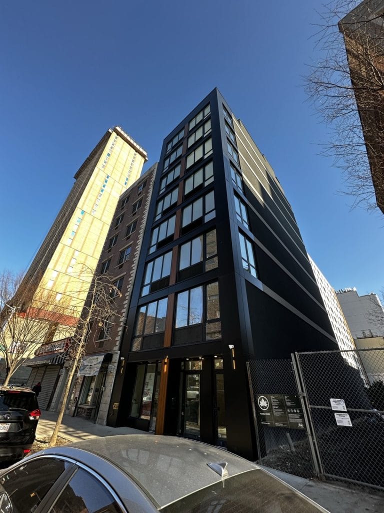 A modern, multi-story building with a sleek black exterior and numerous windows is nestled between two other structures on a bustling city street. A tree stands in front, while to the right lies a fenced-off construction area. Cars are parked neatly along the curb, adding to the urban vibe.