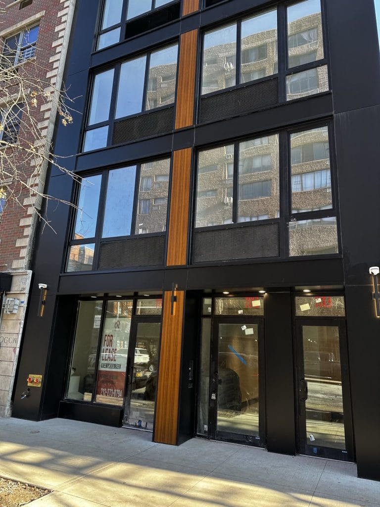 A modern, multi-story building with a black and brown facade stands on a city street. Large glass windows cover the front. A "For Lease" sign is displayed in the ground floor window, providing a contact number for inquiries. Trees are visible on the sidewalk, adding a touch of nature to the sleek architecture.