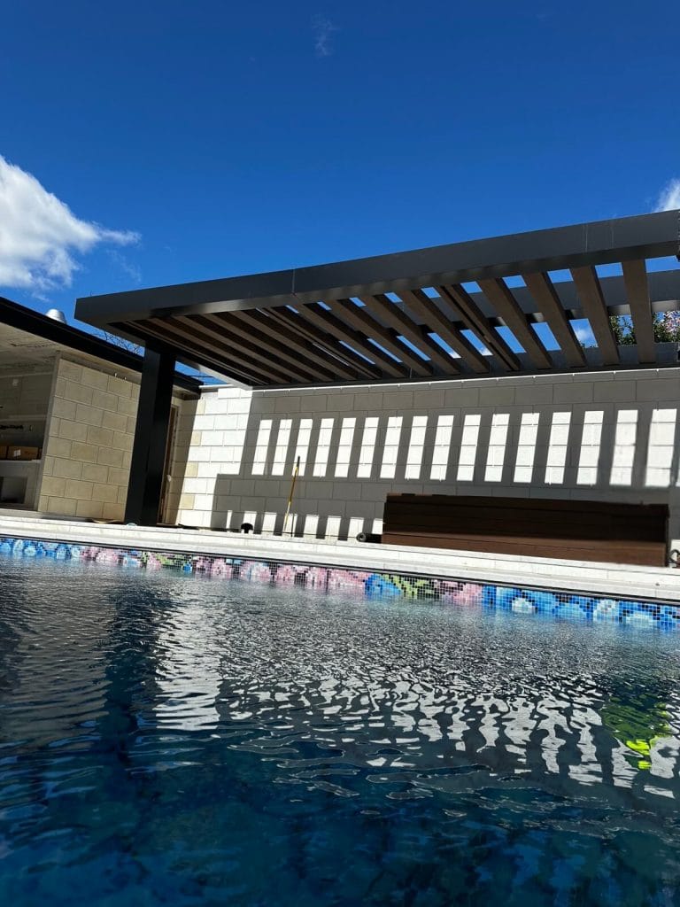 A modern outdoor seating area with a pergola overlooks a sparkling pool adorned with colorful mosaic tiles. The clear, blue sky hints at a perfect sunny day.