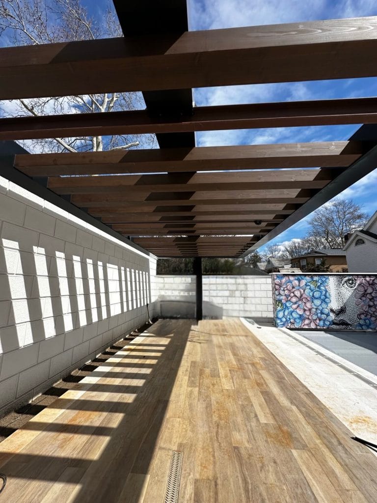 A modern outdoor patio featuring a wooden pergola casting geometric shadows on the light wood floor. To the right is a vibrant mural on a concrete wall, and trees are visible in the background against a clear blue sky. Incorporating eco-friendly design elements, it's a perfect relaxation spot.