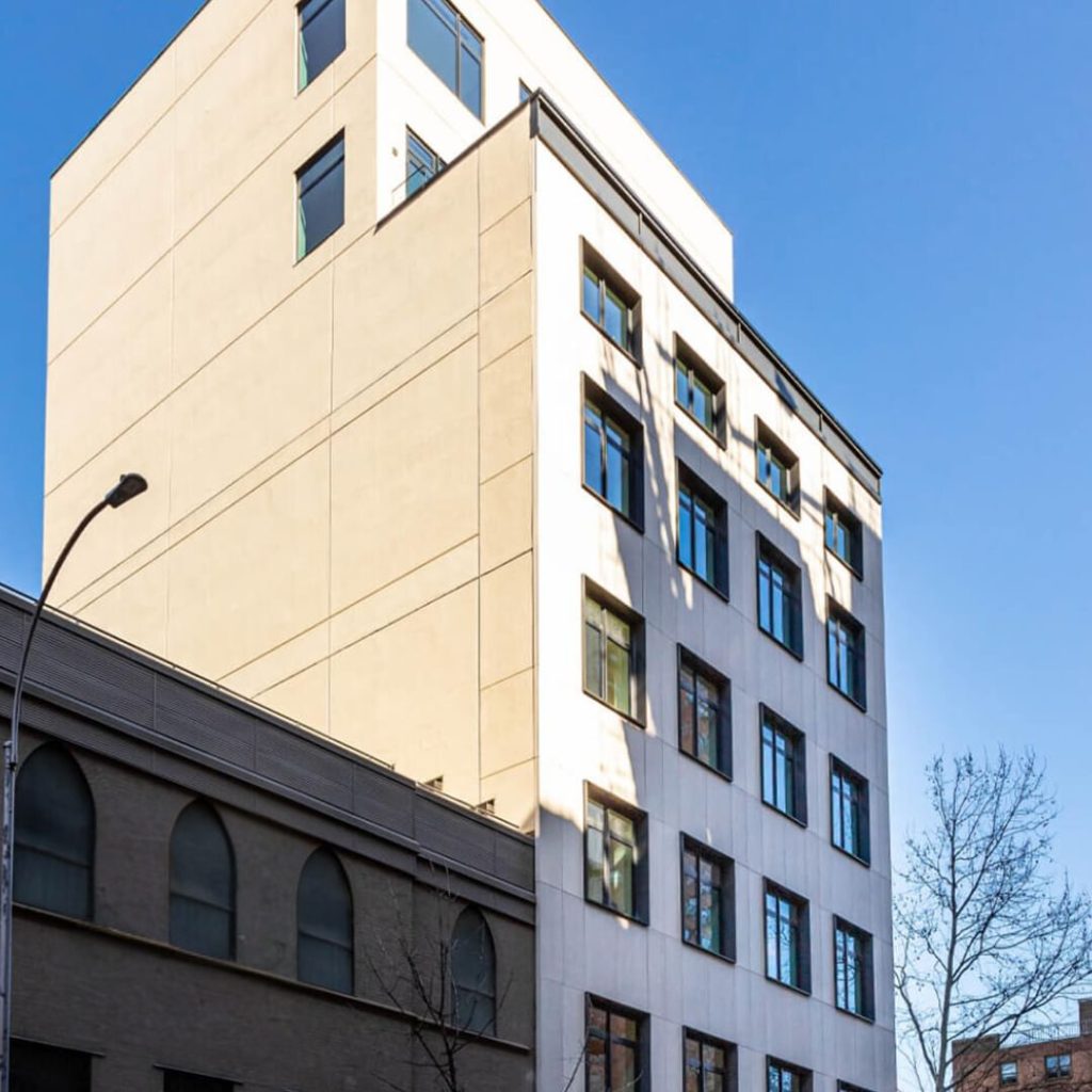 A multi-story building with a modern design features numerous rectangular windows along its facade. The sleek structure, made of concrete and glass, stands against a clear blue sky. In the foreground, a streetlamp and tree branches add to the urban setting.