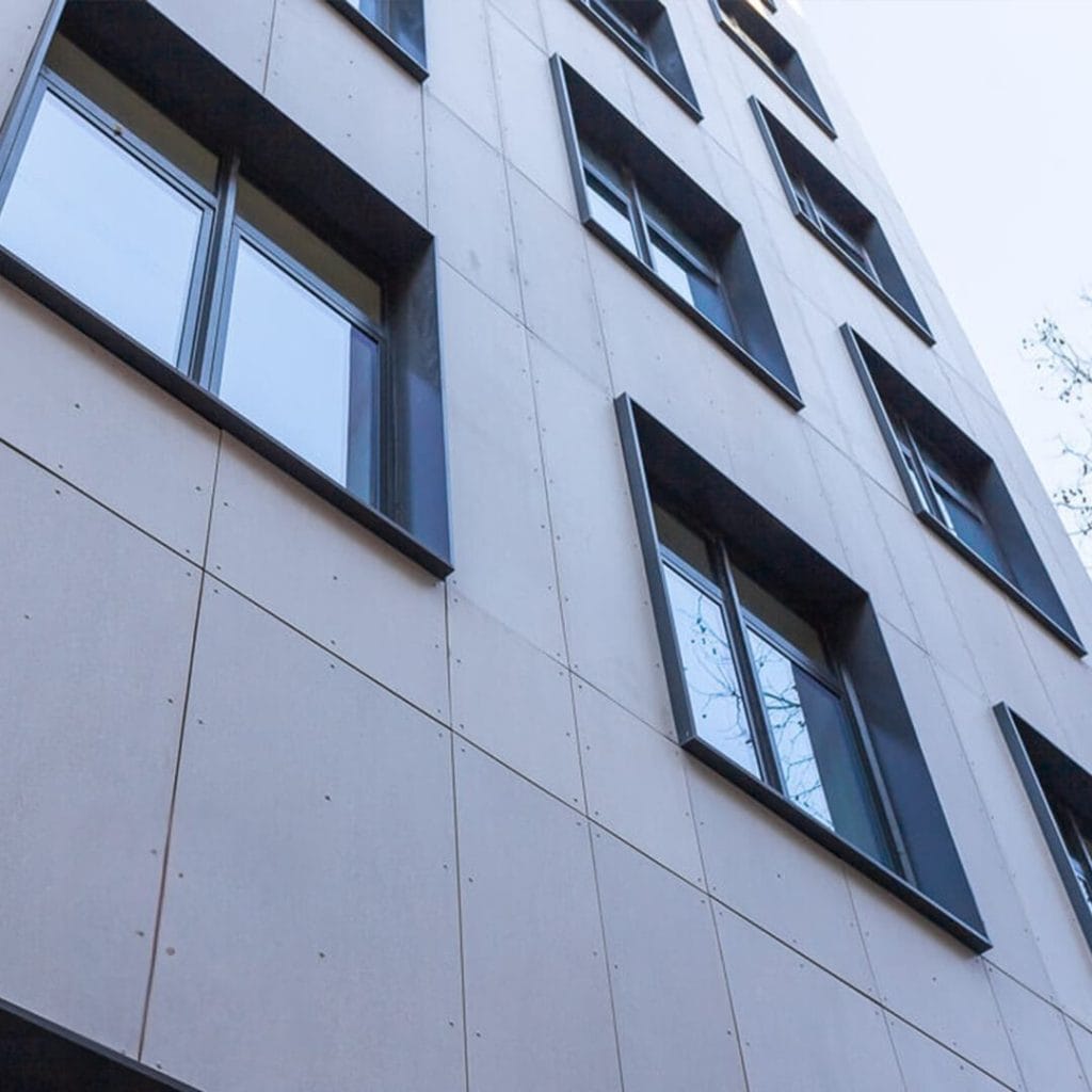 A close-up perspective of a modern building with a beige exterior and large, recessed windows. The angular shadows cast by the window recesses create an abstract geometric pattern on the facade. The clear sky is visible at the top right corner.