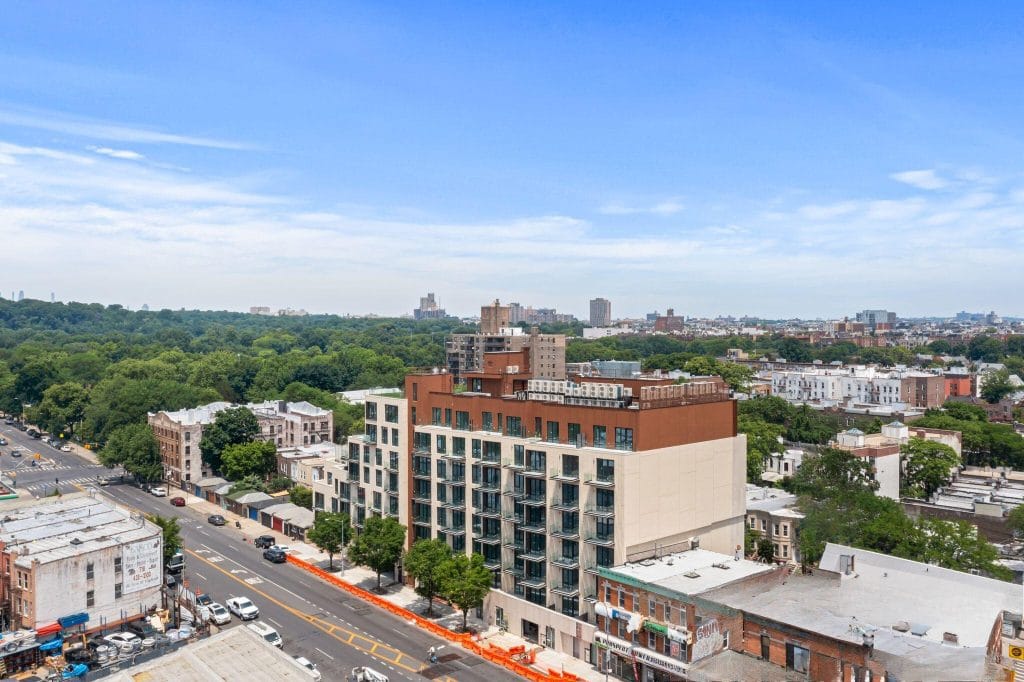 An aerial view of a city neighborhood showcases a modern multi-story apartment building with large balconies. The building is surrounded by smaller structures and tree-lined streets, with a sprawling green park visible in the background under a clear blue sky, presenting an idyllic urban setting.