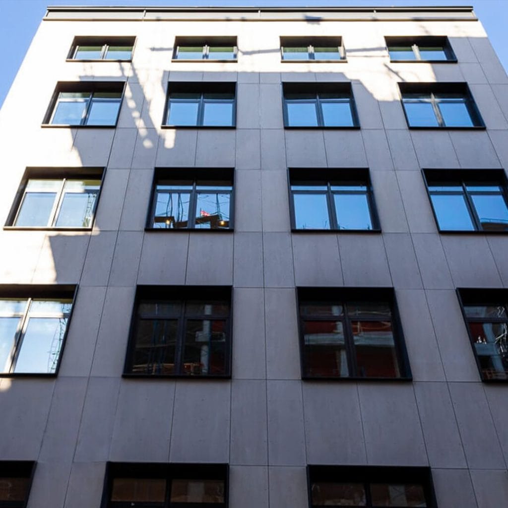 A six-story building with a modern, minimalist facade featuring numerous square windows. The image is taken from the ground level, looking up, highlighting the sleek design and geometric precision of the structure with shadows of nearby structures casting interesting patterns on its surface.
