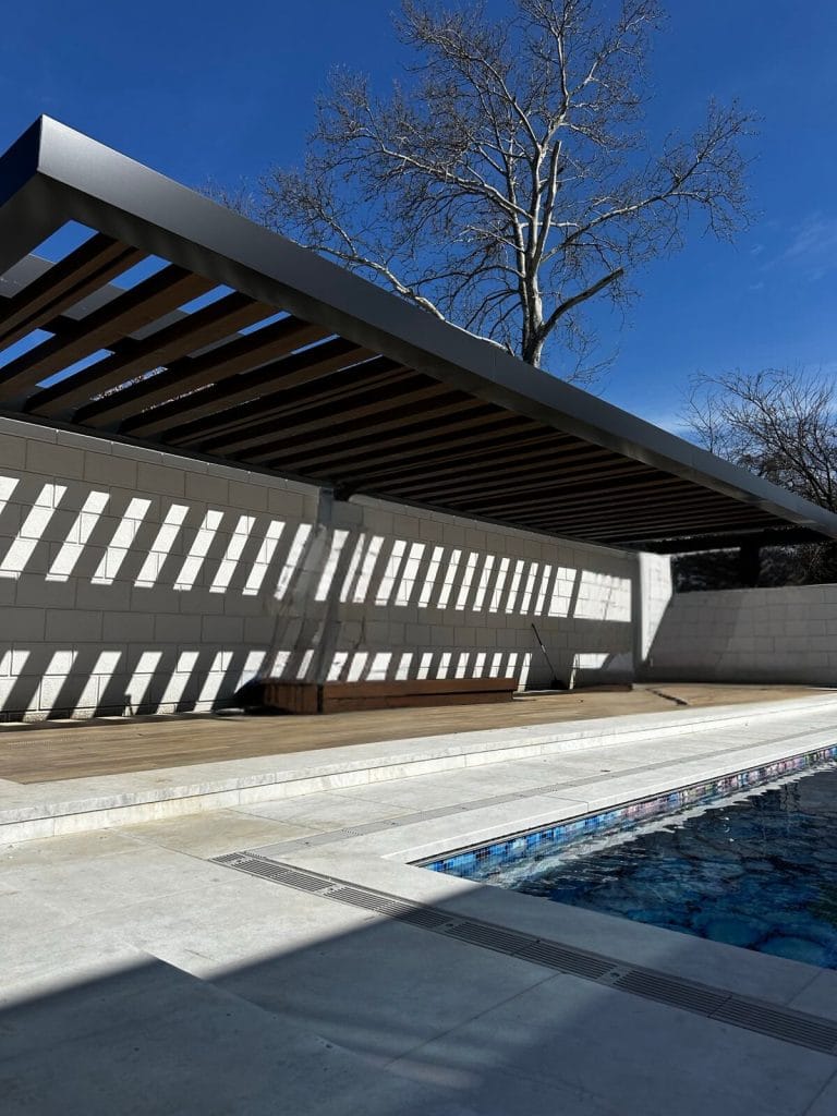 A patio with sleek modern design features a wooden pergola casting striped shadows on the white concrete floor. A bench sits underneath the pergola, and there’s a sparkling swimming pool with intricate tile patterns in the foreground. A leafless tree stands gracefully in the background for a touch of natural elegance.