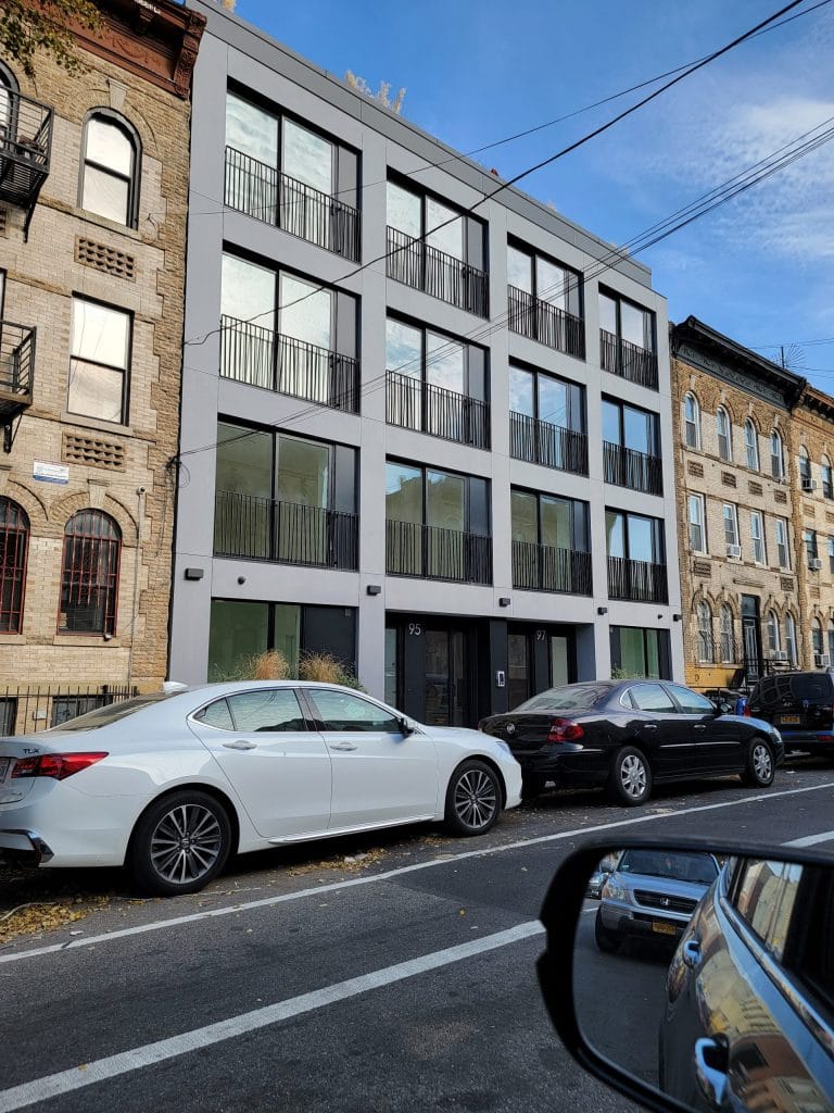 A modern four-story apartment building with large windows and balcony railings stands elegantly, flanked by older, ornate brick buildings. Two cars, one white and one black, are parked neatly on the street in front.
