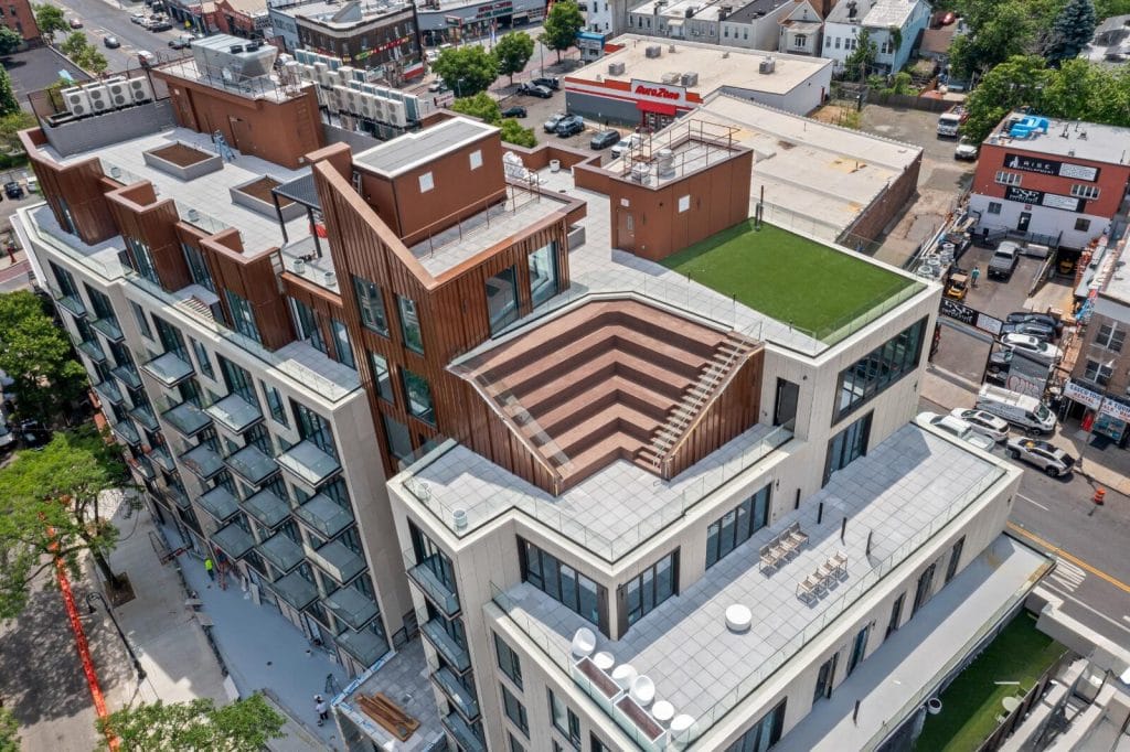 An aerial view showcases a modern, multi-story building with unique architectural features, including a tiered rooftop terrace with seating and a green roof. Surrounding the building are various commercial and residential structures, vibrant streets, and parked cars.