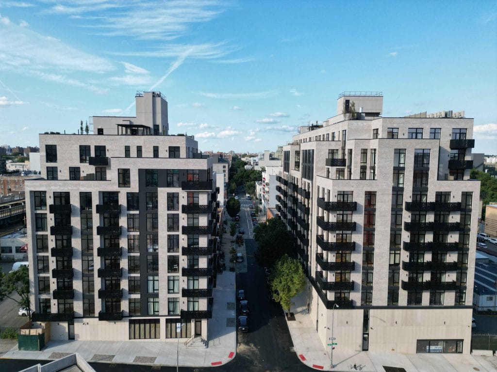 Aerial view of two modern apartment buildings with balconies, symmetrically lining a narrow road. The sky is clear with a few clouds, and additional buildings and streets are visible in the background, offering a picturesque urban scene.