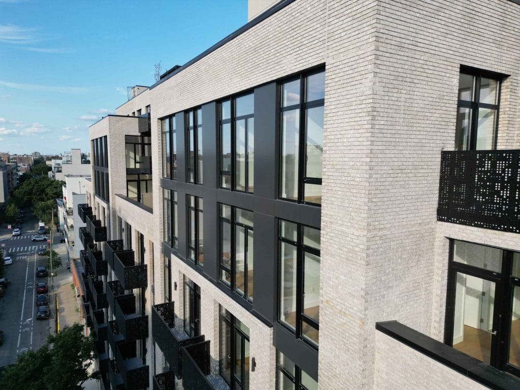 A modern apartment building with a light brick exterior and large, dark-framed windows. Several sections feature intricate, dark-colored balcony railings. A tree-lined street extends towards the horizon under a vibrant night sky, showcasing serene urban living.