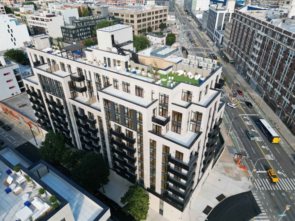 Aerial view of a modern, multi-story apartment building with large windows, balconies, and a rooftop garden. The building is situated at a busy intersection in an urban area, surrounded by other buildings and roads, with some vehicles visible below.
