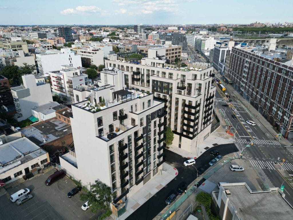 Aerial view of a city block featuring modern mid-rise apartment buildings with balconies and rooftop gardens. Parked cars line the surrounding streets with light traffic. Other residential and commercial structures are visible in the background, adding to the bustling urban vibe.