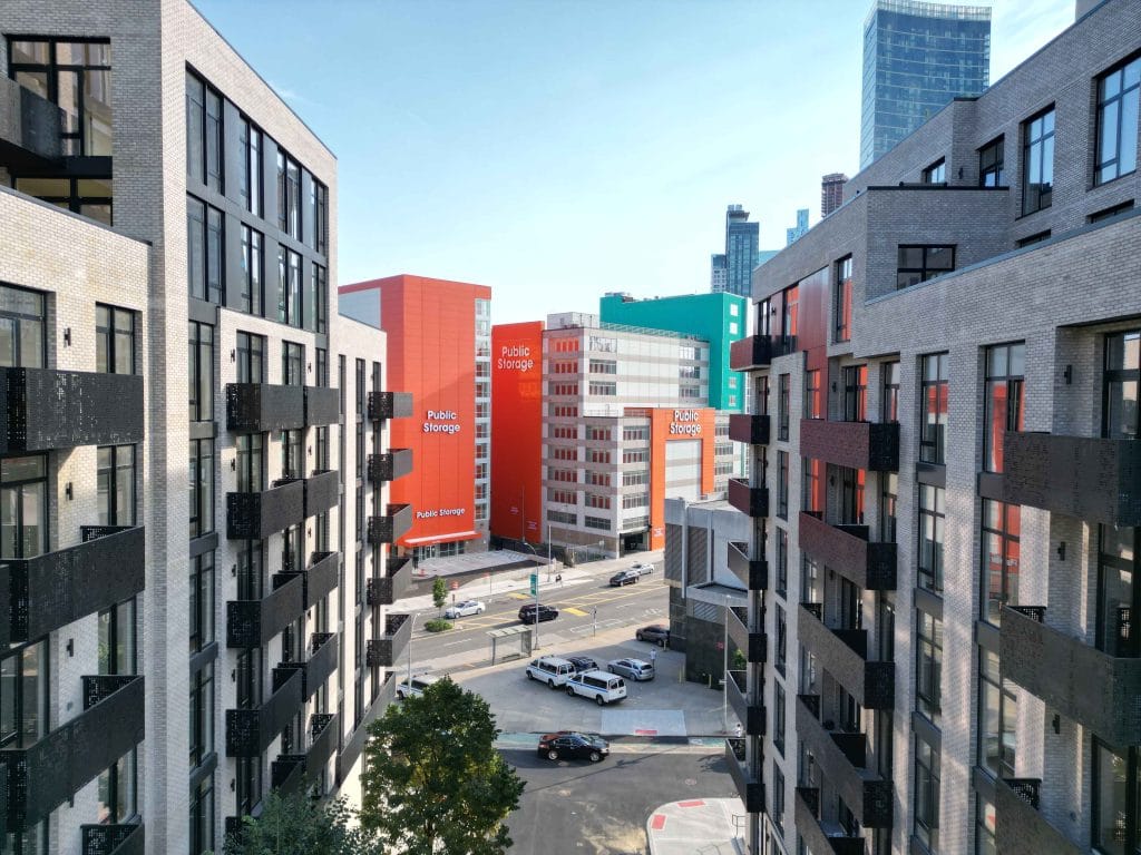 View of an urban street with modern apartment buildings on both sides. In the background, several brightly colored buildings with signs for "Public Storage" can be seen. A few vehicles are visible on the street, and there are trees and pedestrians going about their day, giving the area a lively feel.
