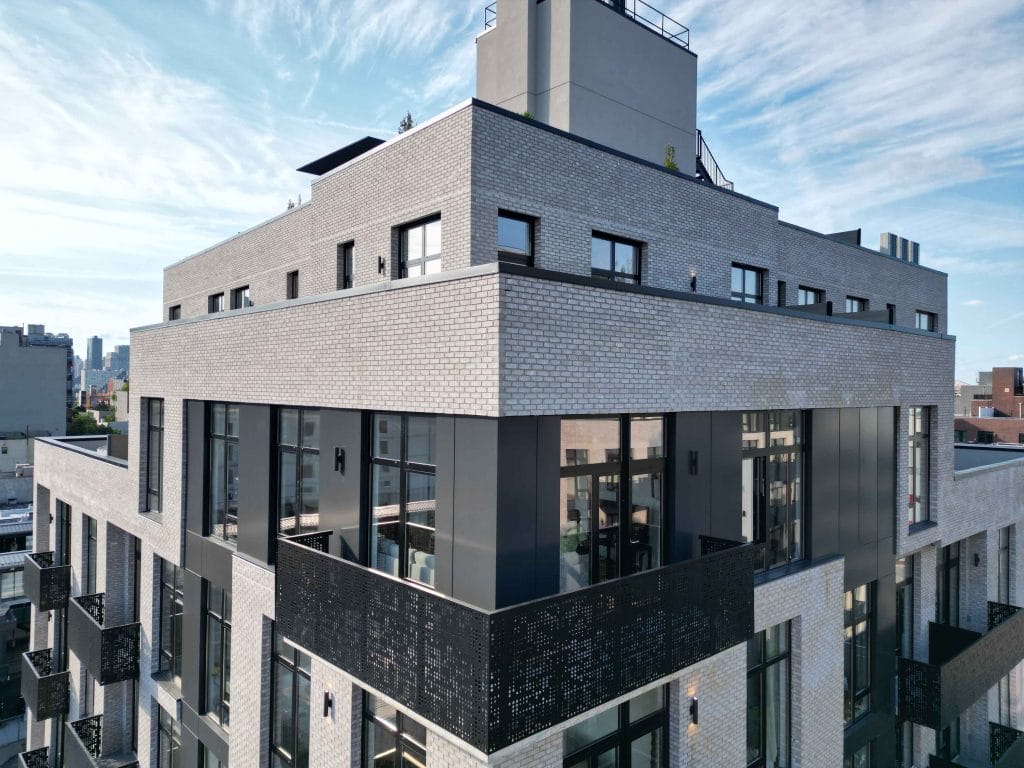 A modern, multi-story building with a contemporary design. The exterior features a mix of light-colored bricks and large, dark-framed windows. Balconies with black railings are visible, and the top of the building boasts a stylish rooftop area against a partly cloudy sky.