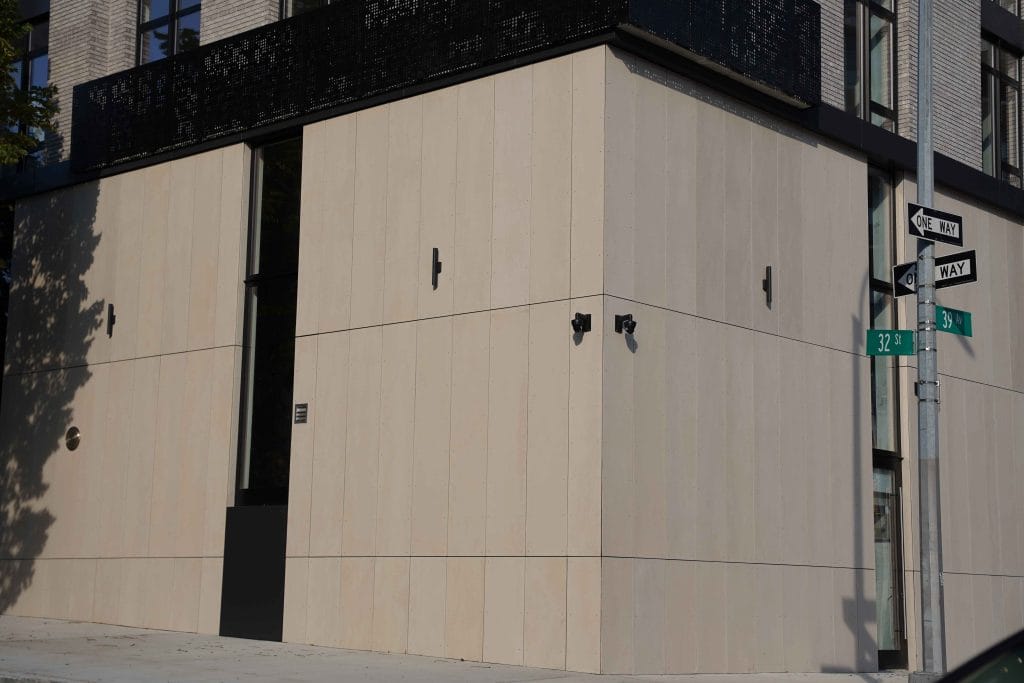A contemporary, beige building with black accents is situated at the intersection of 32nd Street and 10th Avenue. The corner of the structure features a rectangular window and two security cameras. Street signs are visible on the right side of the image, enhancing its urban feel.