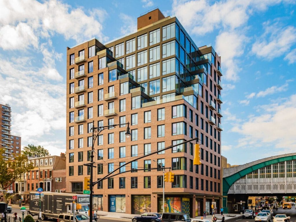 A sleek multi-story apartment building with a mix of brick and glass exterior stands on a bustling urban street corner. Cars and trucks drive by, pedestrians stroll on the sidewalks, and a clear blue sky with scattered clouds is overhead, highlighting the vibrant city atmosphere.