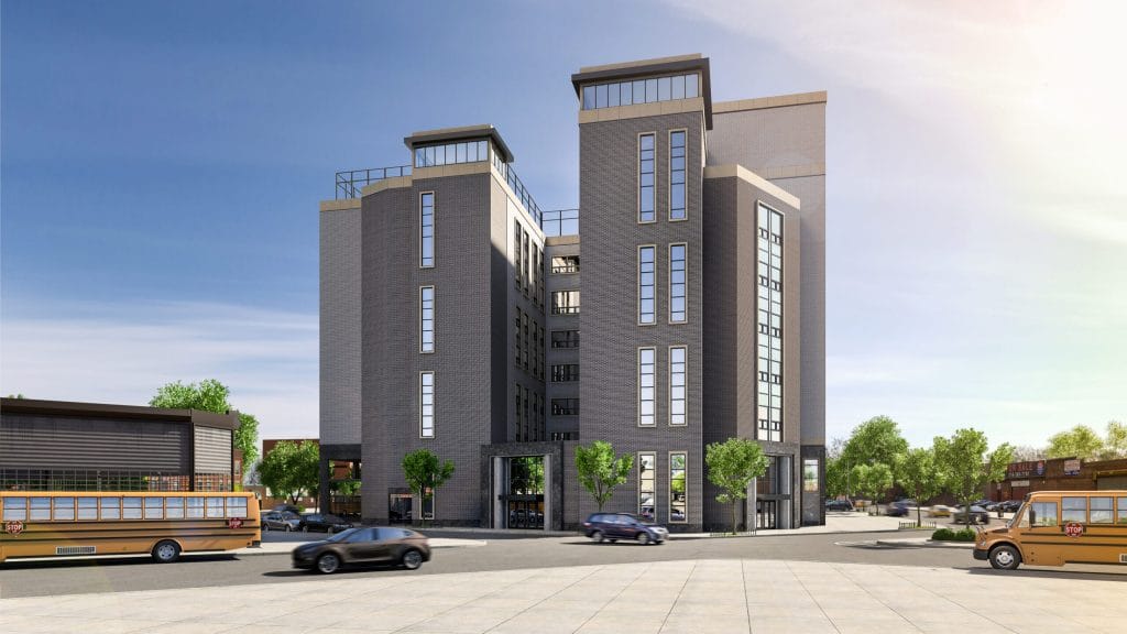 A modern, mid-rise building with gray brick and large windows stands against a clear blue sky. Two school buses and several cars are on the tree-lined street surrounding the building, with smaller buildings visible in the background.
