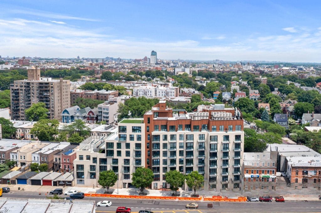 Aerial view of a cityscape featuring a mid-rise residential building in the foreground with a mix of older buildings, trees, and various unique architectural styles in the background. The landscape extends towards the horizon under a clear blue sky.
