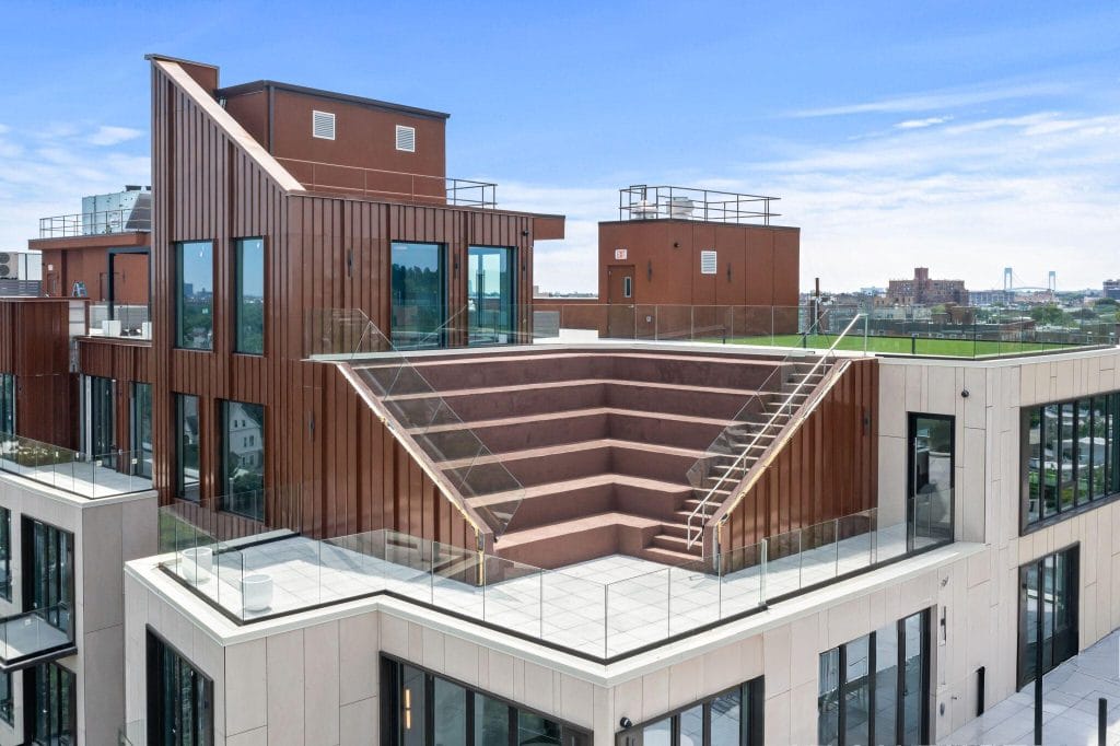 A modern, multi-story rooftop with unique architecture featuring angular design elements, a gray and white exterior, expansive glass windows, tiered platforms, and glass railings. The background shows a cityscape under a clear blue sky.