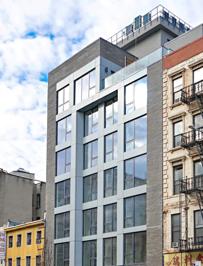 A modern multi-story building with large reflective windows and a rooftop structure stands between older buildings with diverse facades. The sky is partly cloudy, adding to the city's dynamic charm.