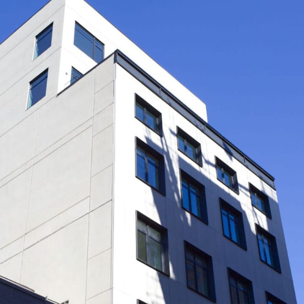 A modern, multi-story building with large windows stands against a clear, blue sky. Made of concrete or a similar material, it features clean lines and a minimalist design. Shadows from nearby structures cast intriguing patterns on its facade.