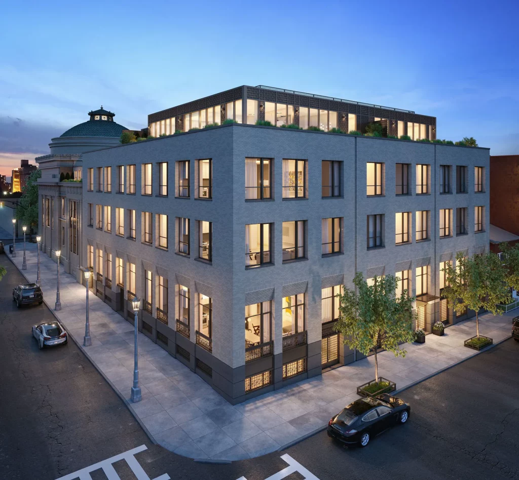 A modern four-story apartment building on a street corner at dusk, showcasing large windows with warm lighting inside. The building features a rooftop terrace and small trees along the sidewalk. Nearby cars are parked alongside the building and on adjacent streets, adding to the urban charm.