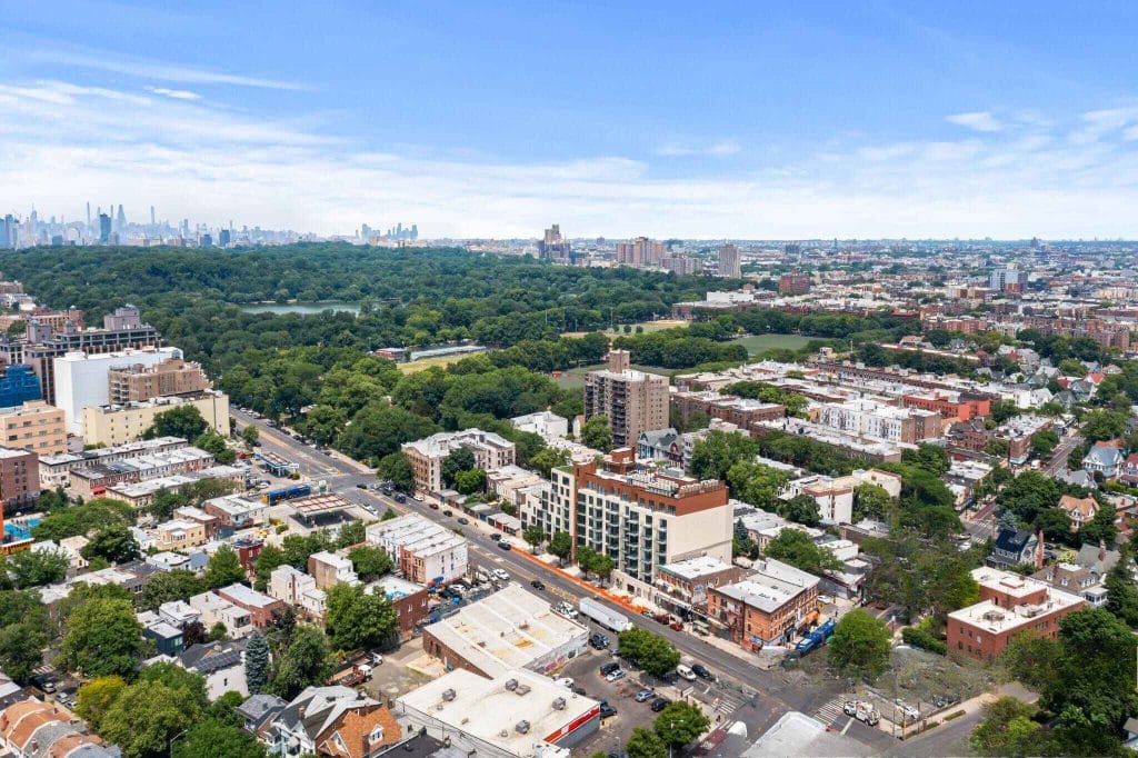 Aerial view of a cityscape with lush green parks and a mix of tall buildings and houses. A distant metropolitan skyline under a clear blue sky enhances the vista, while roads and parked cars add to the dynamic urban scenery.