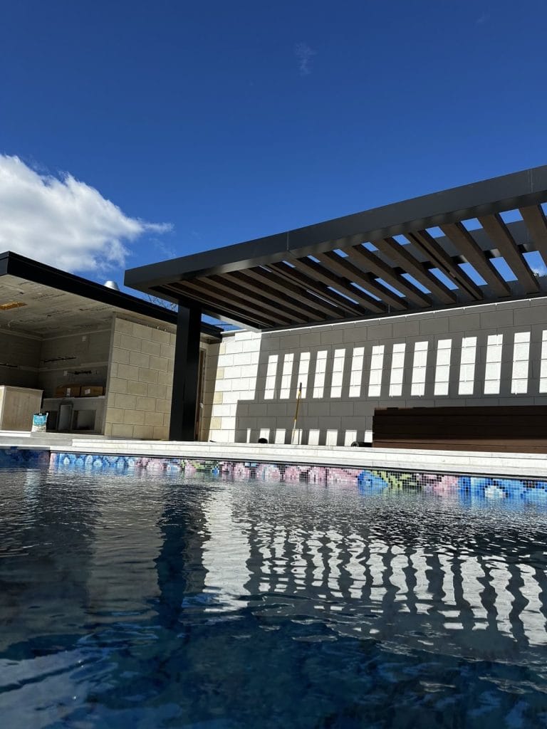 A modern outdoor pool features clear blue water with a pergola providing shade over a nearby seating area. Under the pergola, there is a wooden bench and an elegant kitchen area against a stone wall. The sky is clear with scattered clouds enhancing the serene ambiance.