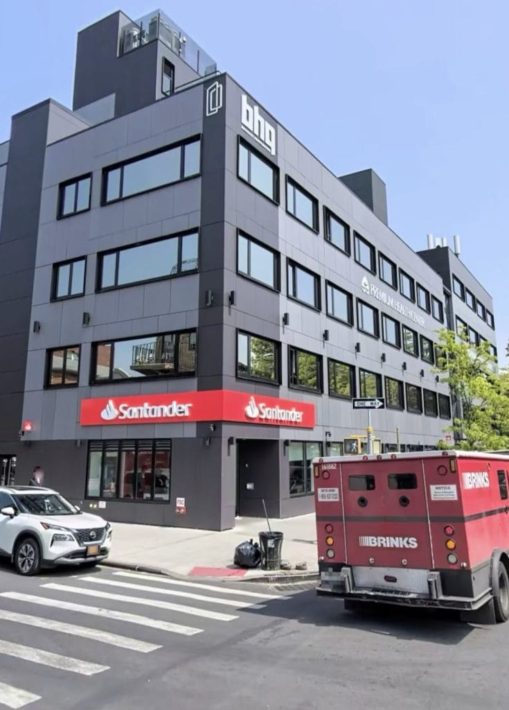 A multi-story black building with Santander and BGC logos on the facade. Ground floor windows display the Santander logo with a red sign. A Brinks armored truck is parked on the street next to a silver SUV and a pedestrian, all under a clear, sunny sky.