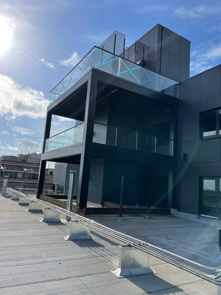 A modern rooftop view showcases a two-story black structure with glass railings and large windows. The clear and sunny sky highlights its architectural details, with nearby rooftops visible in the background.