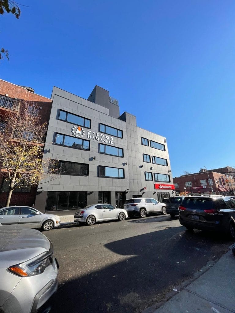 A four-story gray building against a clear blue sky, with "Ramada" and its logo displayed on the facade. Ground-level shops, including a cozy café and a Vodafone store, line the street with parked cars in front. Adjacent buildings are partially visible.